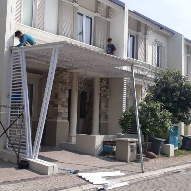 Pemasangan kanopi PVC putih di teras rumah bergaya klasik, dengan dua pekerja menyelesaikan struktur kanopi tahan panas dan hujan.