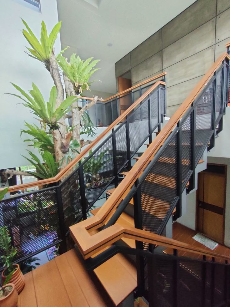 Tangga besi dengan anak tangga kayu Tangga besi rumah bertingkat minimalis dengan desain modern, memadukan pijakan kayu dan railing besi hitam berlubang, dikelilingi tanaman hias tropis di area interior bernuansa industrial.