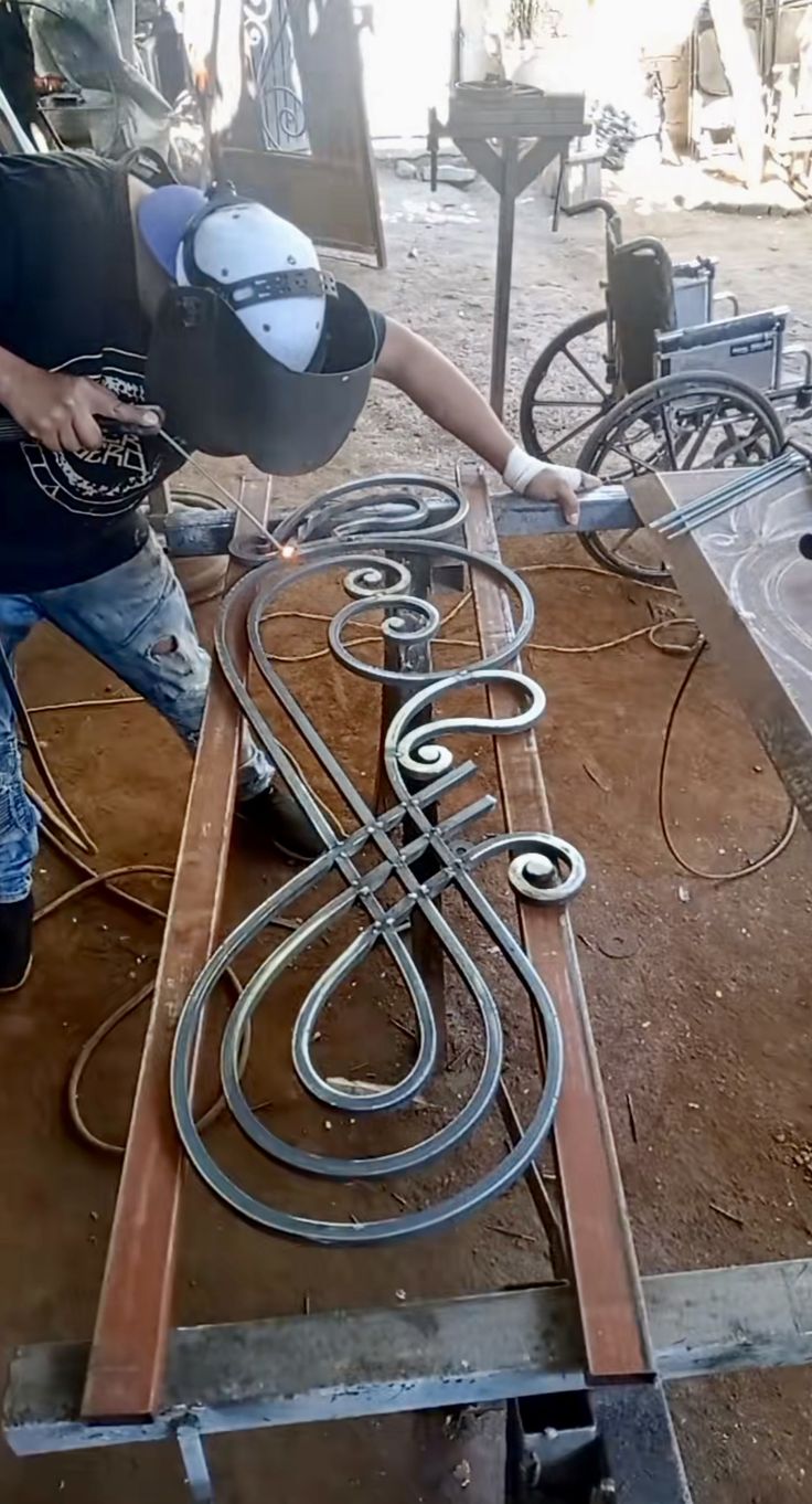 Tukang las sedang mengerjakan desain teralis besi artistik dengan motif lengkung dan ornamen klasik di bengkel las manual.