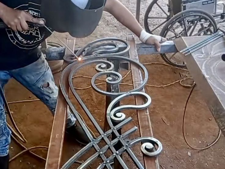 Tukang las sedang mengerjakan desain teralis besi artistik dengan motif lengkung dan ornamen klasik di bengkel las manual.