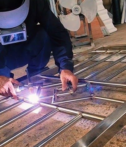 Tukang las profesional sedang mengelas rangka pagar besi stainless dengan presisi di bengkel las, menggunakan pelindung wajah dan alat las listrik.