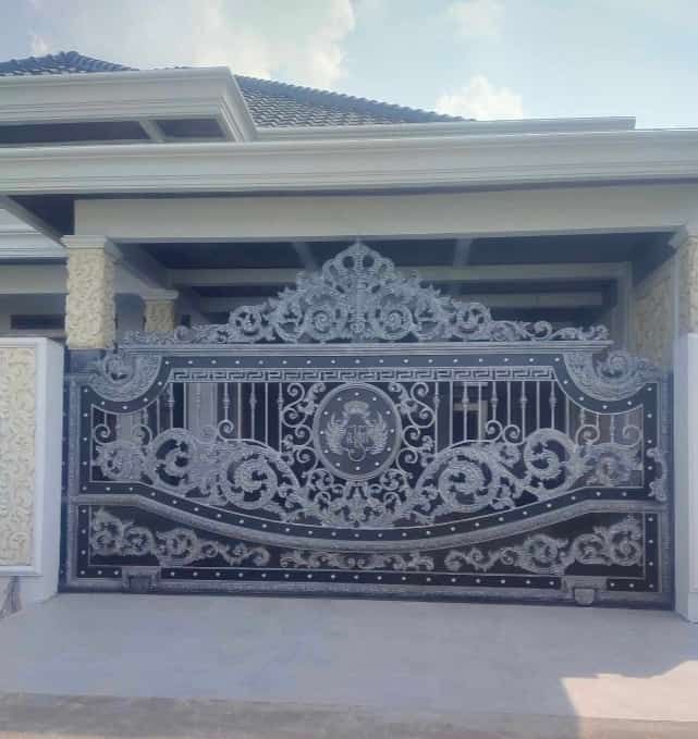 Pintu pagar besi tempa mewah dengan ornamen klasik warna hitam dan silver di depan rumah bergaya modern tropis.