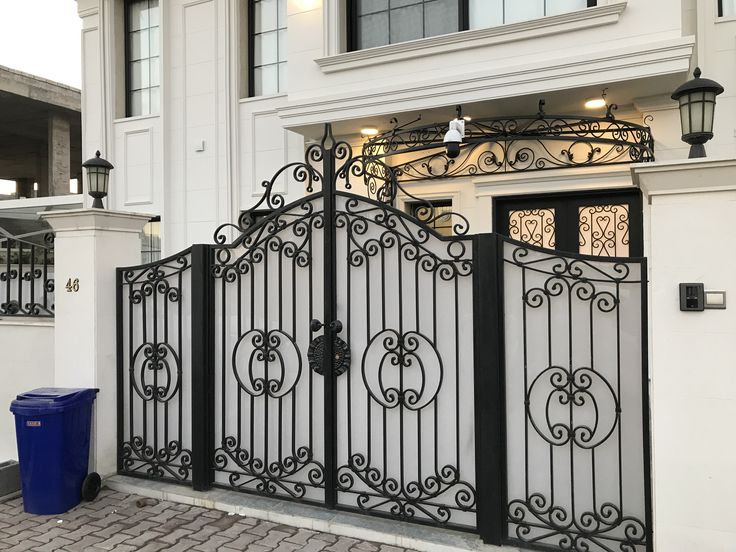 Pagar besi tempa klasik berwarna hitam dengan desain lengkung dan ornamen elegan, terpasang di depan rumah mewah bergaya Eropa modern.